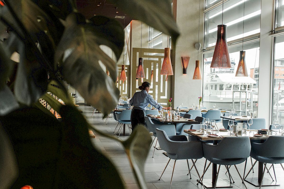 A modern restaurant with a person setting tables, stylish chairs, and pendant lights, viewed through large plants by the window.