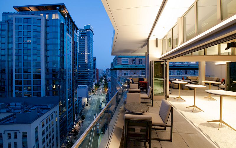 A modern cityscape with tall buildings at dusk, featuring a sleek balcony with tables and chairs overlooking the lively street below.