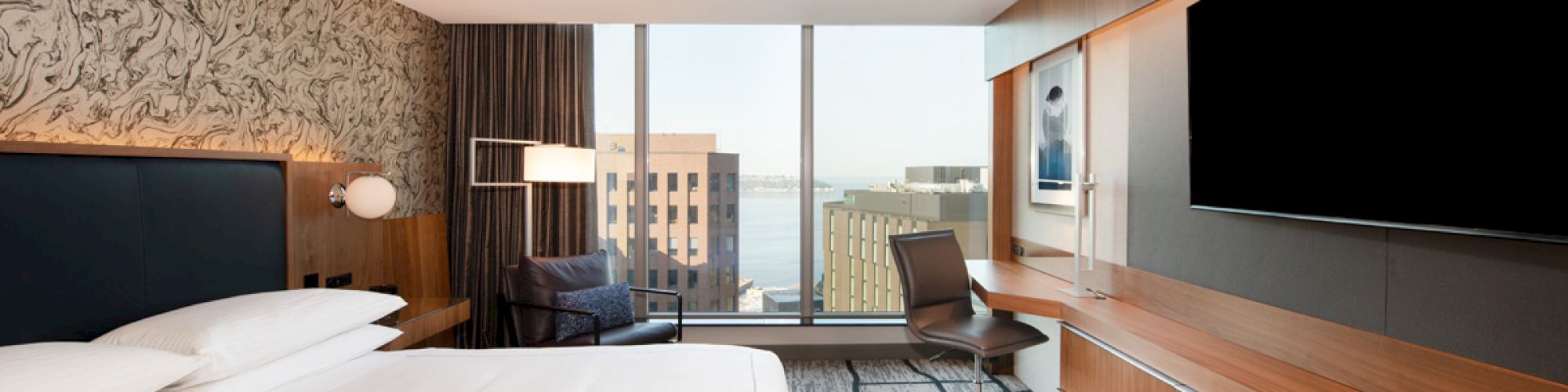 A modern hotel room features a large bed, desk, chair, flat-screen TV, and large window with a city view.